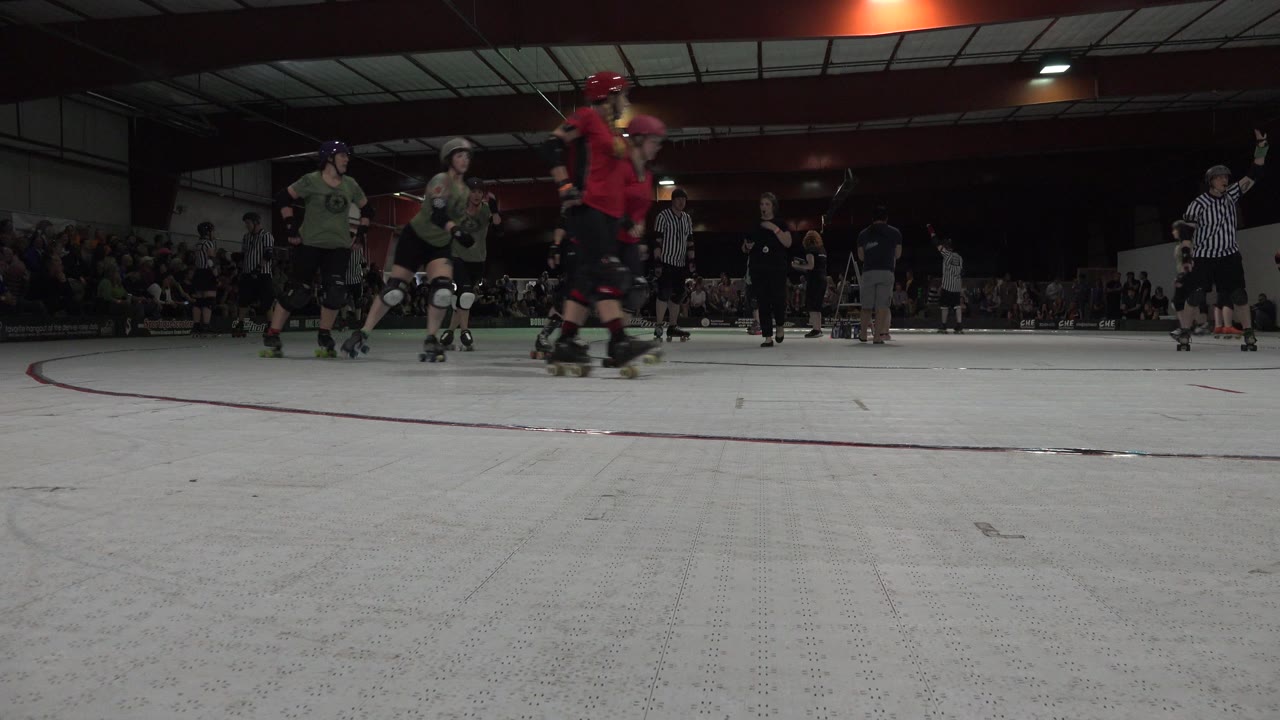 Skaters and referees on an indoor skating track with spectators in the background.