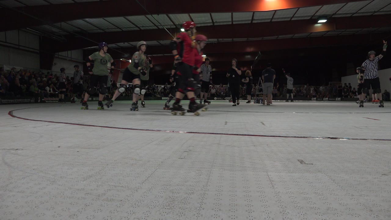 Denver indoor skating scene with skaters in green and red shirts crossing the foreground, officials in striped shirts, and a seated audience in the dim background.