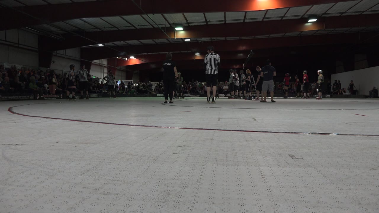 A referee and several skaters stand on the gym floor in front of a crowd.