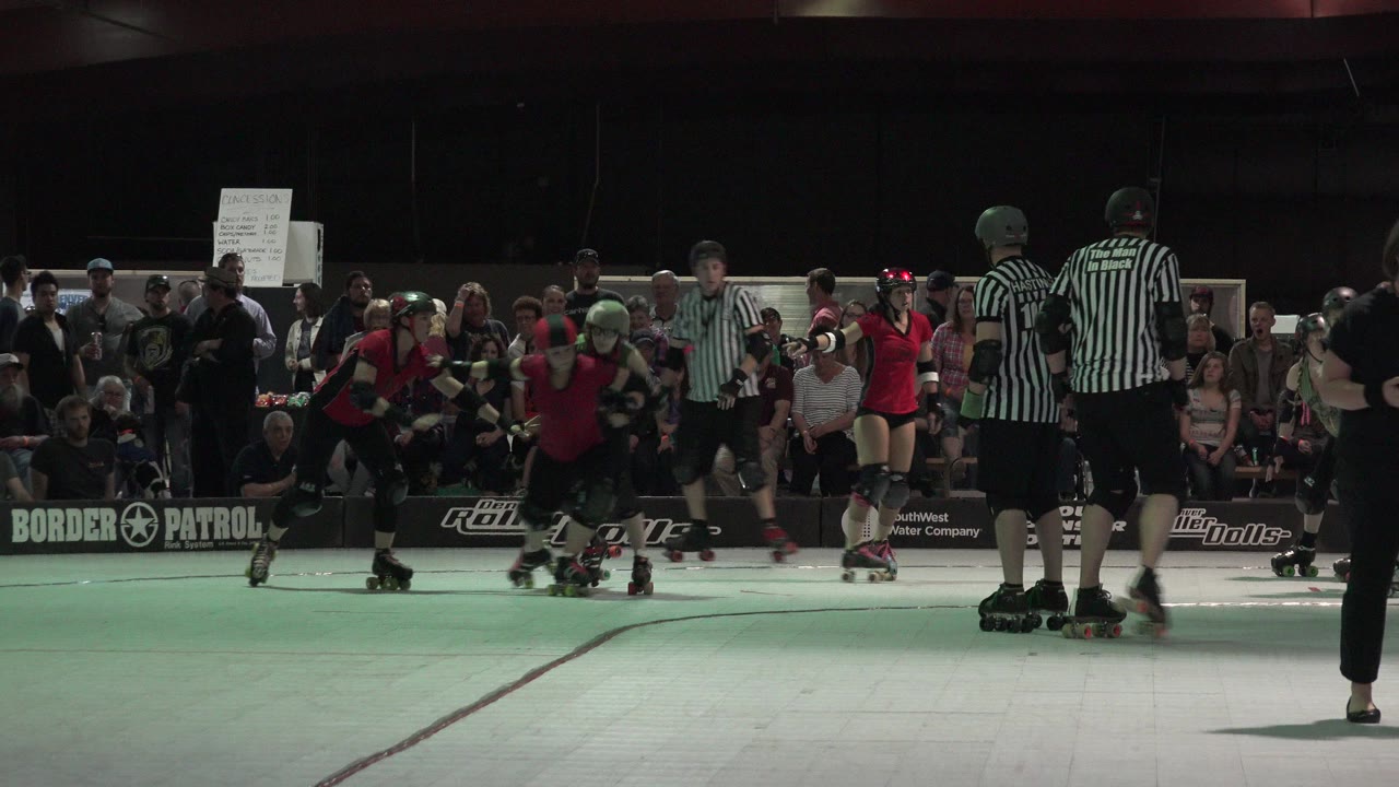 Roller derby skaters in red shirts move past referees and spectators.