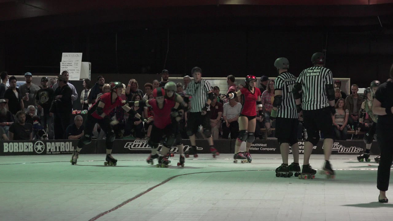 A cluster of women roller derby skaters in red and black gear moves near two referees as spectators watch from the background in Denver, Colorado.