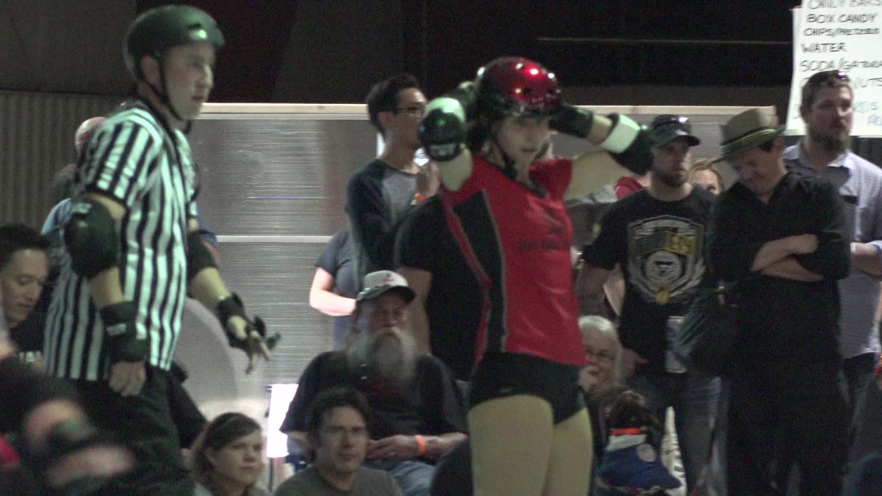 In Denver, Colorado, a red-uniformed skater with a glossy helmet raises an arm near protective pads as a referee and spectators fill the background.