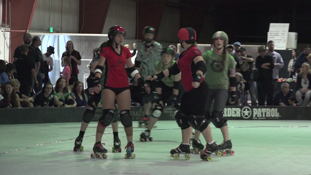 At 0:01 in Denver, Colorado, skaters in helmets and pads pass near each other on the indoor track while the seated audience watches from behind the barrier.