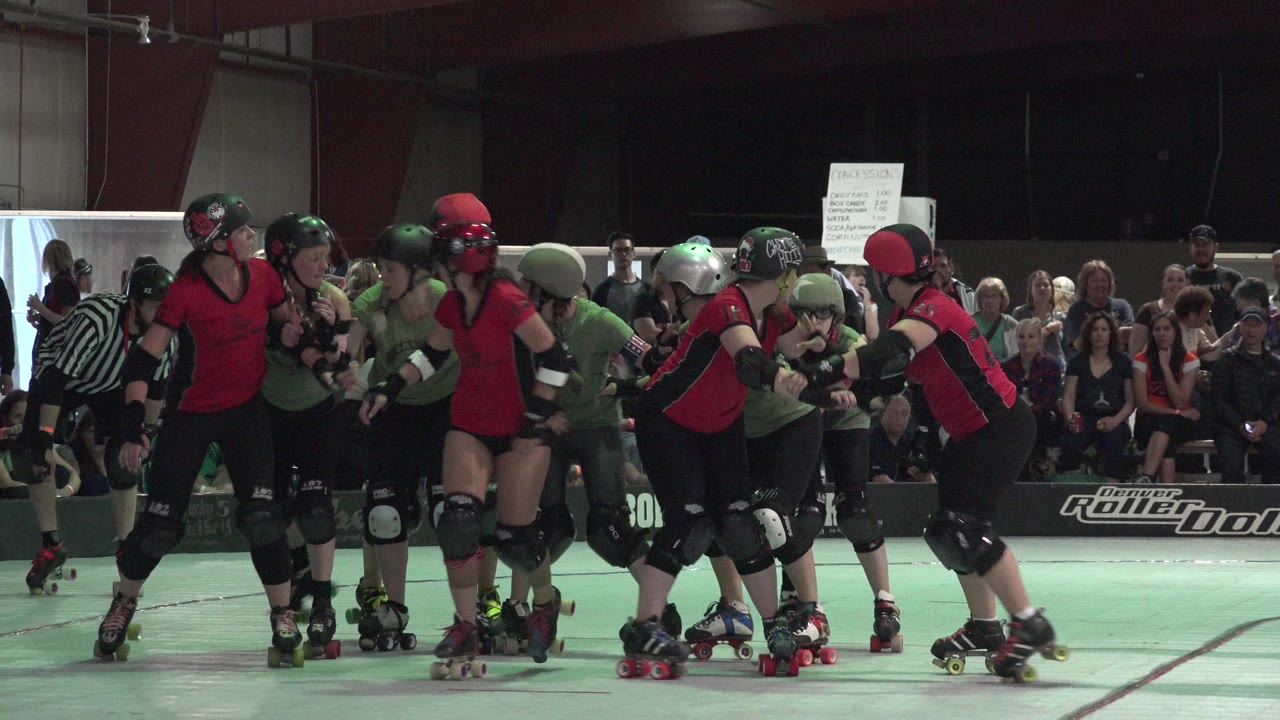 Denver roller derby skaters in red and green uniforms crowd together near a referee, with spectators seated behind the rink barrier.