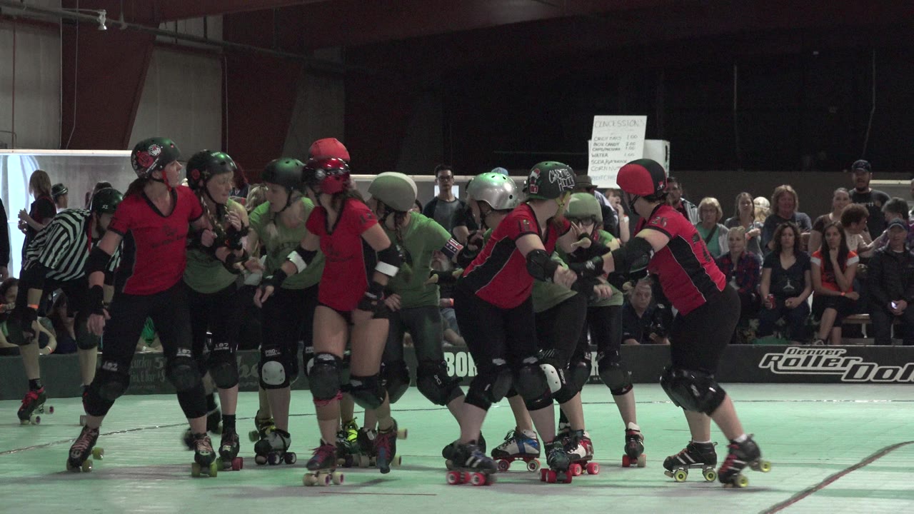 Denver roller derby players lean into contact in a tight pack, with red jerseys, green shirts, helmets, pads, and spectators behind them.