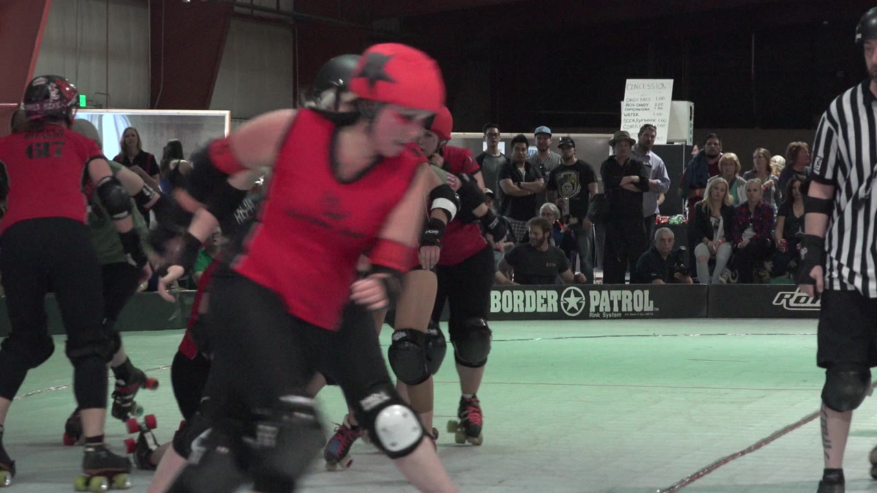 A red-clad skater crosses the foreground in Denver as another teammate and a striped referee stand near the indoor roller rink crowd.