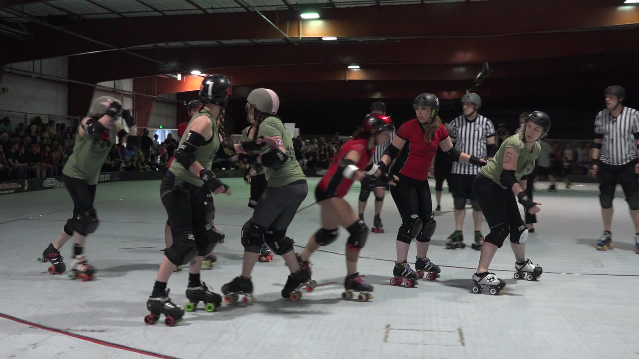 Denver roller derby players in red and green uniforms skate past each other on the white rink while a striped referee watches from the background.