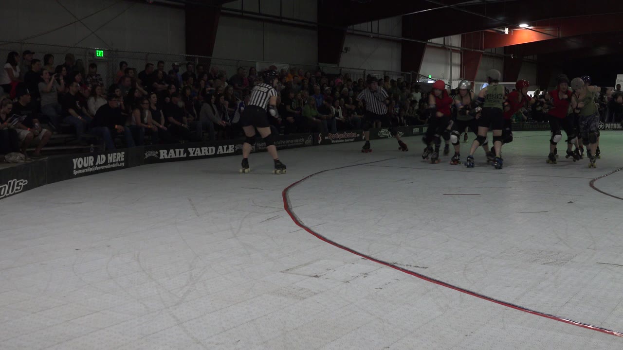 Denver roller derby match viewed from rink level, with skaters and striped referees moving along the far side of the white track as spectators fill the dark stands behind them.