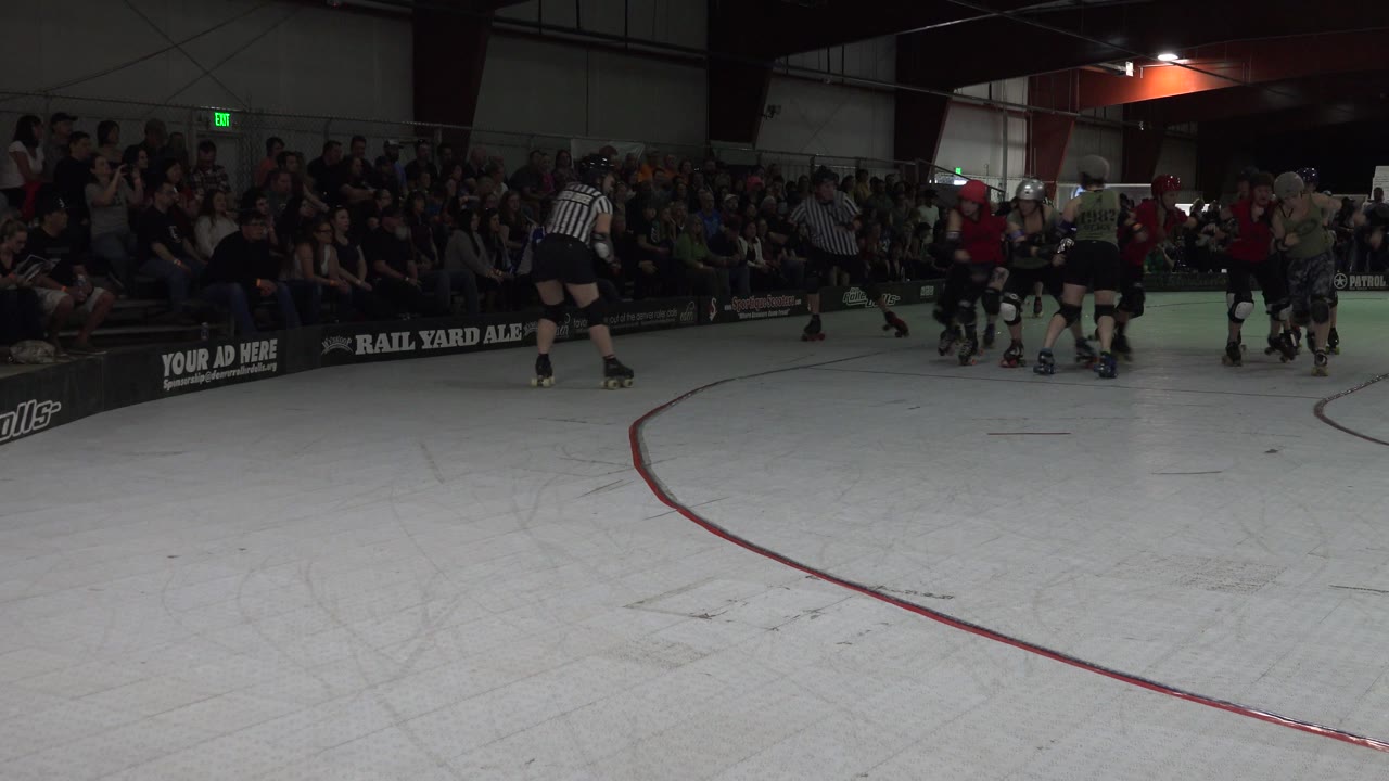 A referee in stripes stands in the foreground area while roller derby skaters cluster farther across the Denver rink, with crowded stands and dark venue walls in the background.