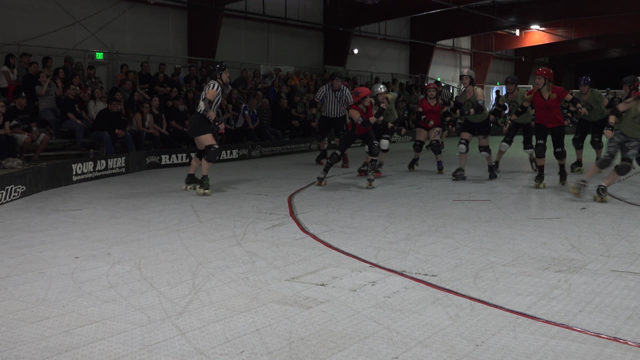 Roller derby skaters pass on the right side of the rink in front of spectators.
