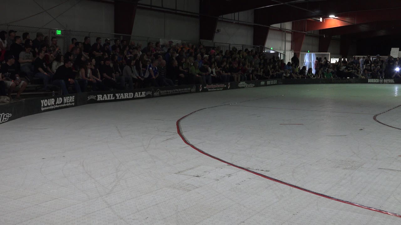 Denver indoor roller rink seen from rink level, with a red curved line on the pale floor, spectators seated in dark bleachers, and no visible skaters in the open foreground.