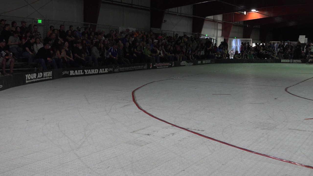 Opening frame of a Denver indoor roller rink with a pale skating surface, red curved line, dark rink boards, advertising signage, and spectators seated along the far side.