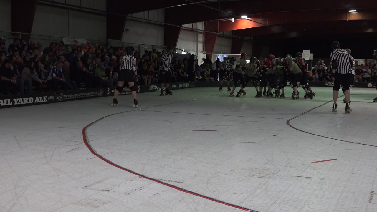 Denver 2015 frame showing skaters bunched near the center of the indoor rink while a referee watches from the right and the audience sits behind the rail.