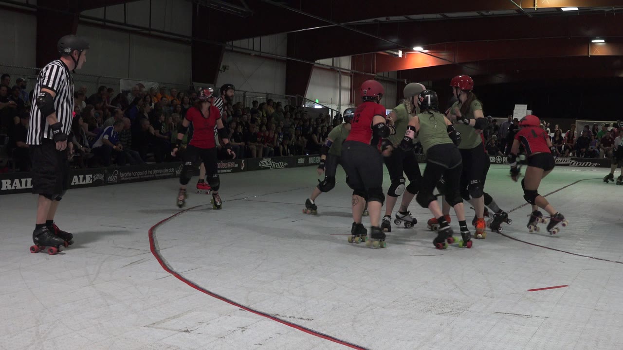 Roller skaters and a referee on an indoor rink with spectators behind them.