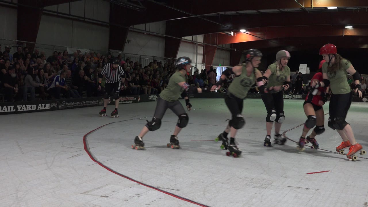 A group of roller derby skaters in green and red uniforms leans into the rink curve in Denver, Colorado, with spectators filling the background bleachers.