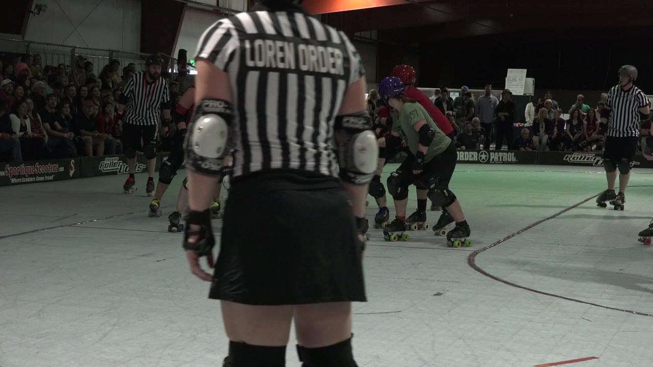 In Denver, Colorado, the referee remains centered in the foreground as derby players skate and cluster behind them under dim indoor arena lighting.