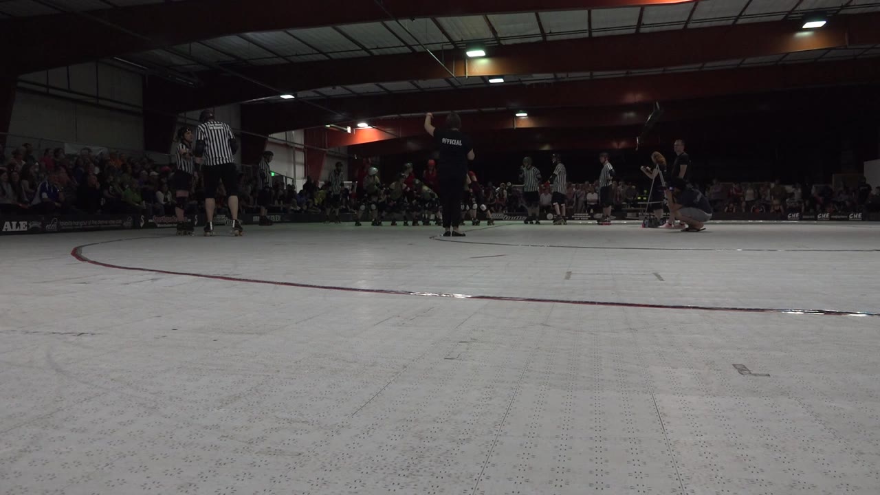 Low ground-camera view across a Denver indoor roller rink, with a striped referee at left, skaters in dark uniforms near the center, and spectators seated along the back wall.