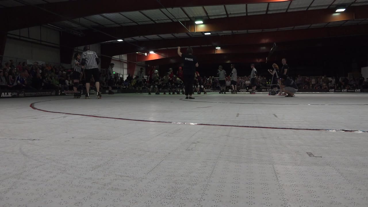 The Denver roller rink remains framed from floor level, with a referee standing at the left edge, skaters gathered near the far side, and the audience visible behind them.