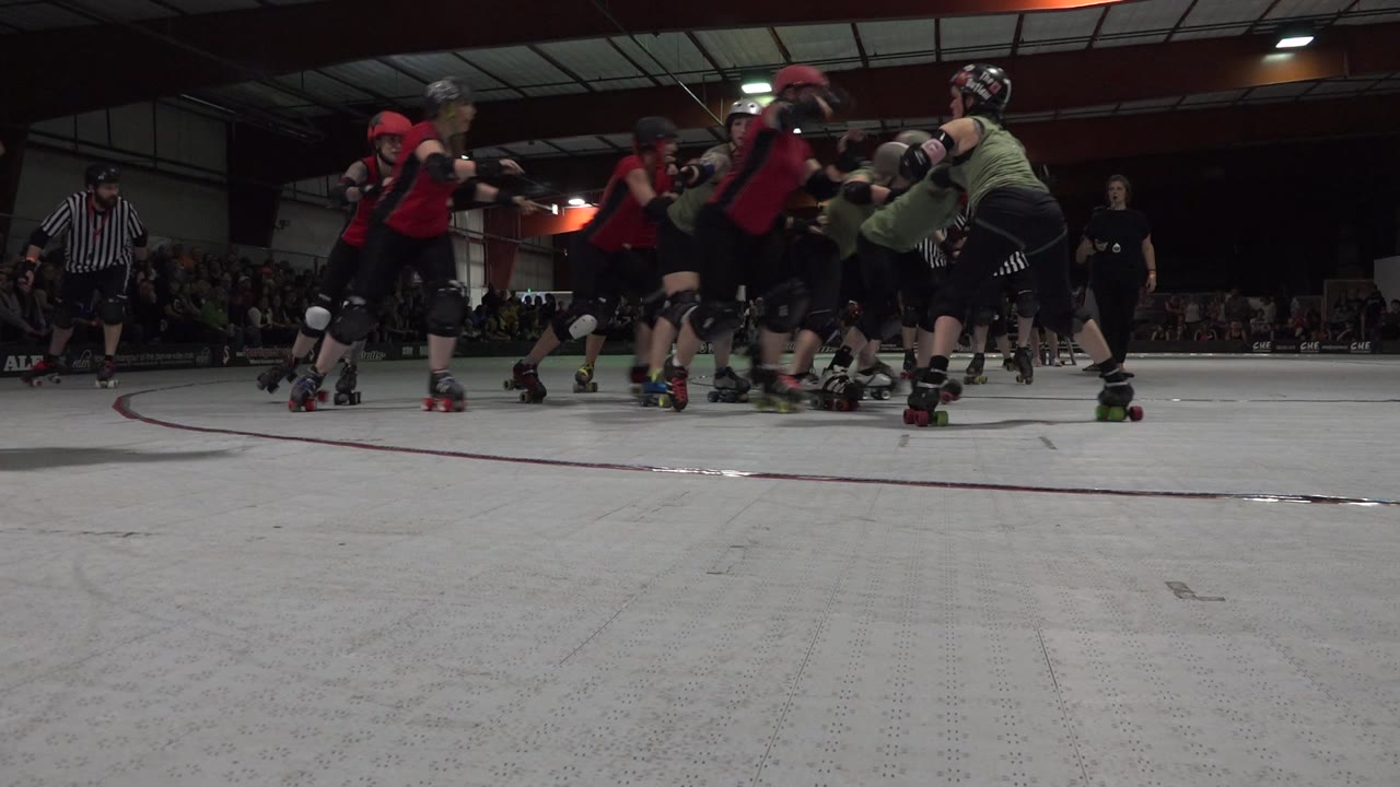 Roller skaters cluster on an indoor rink while spectators watch from the background.