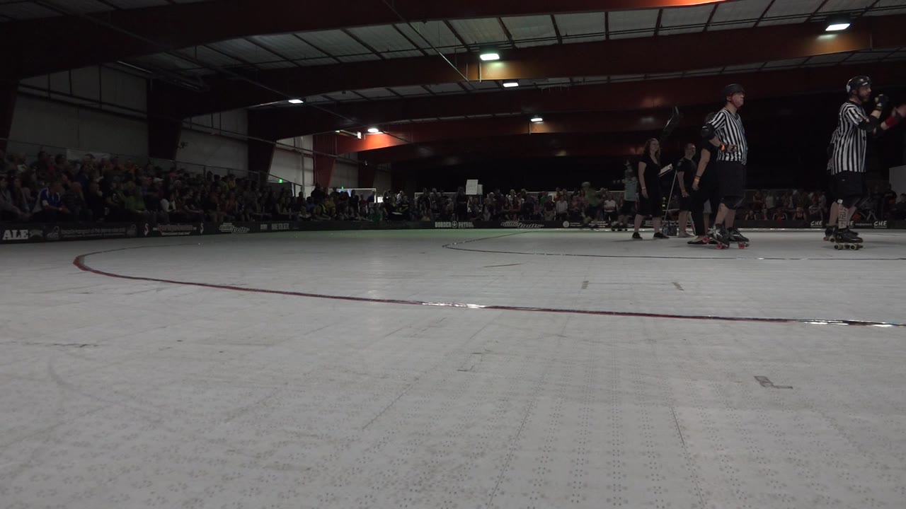 Low-angle view of a Denver, Colorado indoor skating rink with referees and roller skaters at the right edge and a seated crowd across the background.