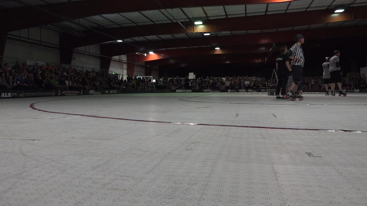 Indoor skating rink with crowd in bleachers and officials at right.