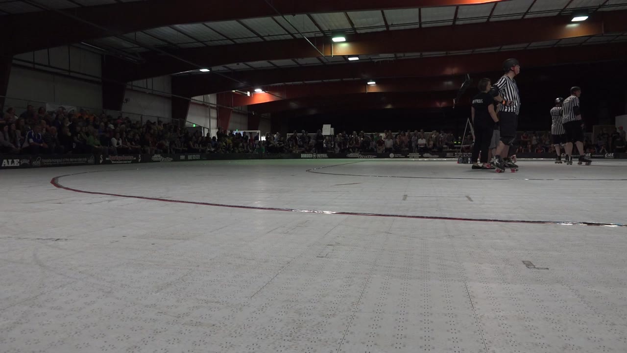 A broad pale skating surface dominates the foreground while Denver event spectators sit in dark bleachers and a small group of officials and skaters remains clustered to the right.