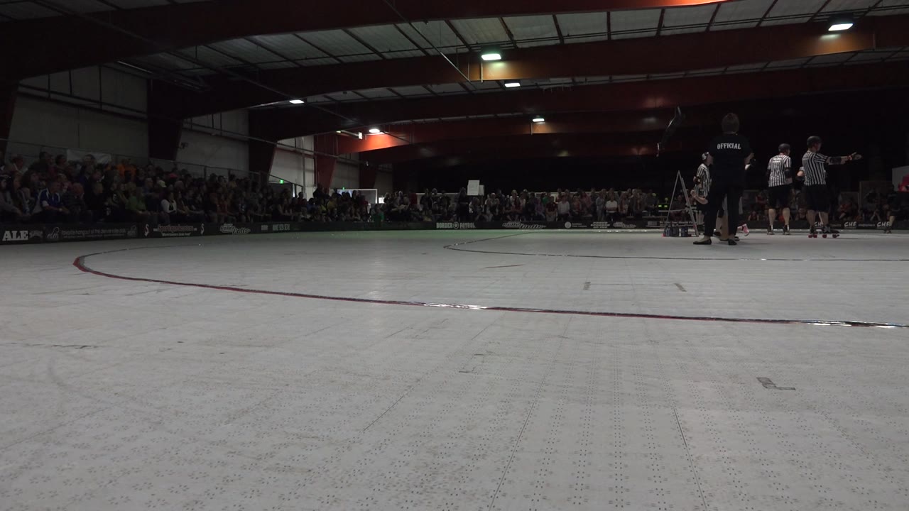Low view of an indoor roller derby rink with referees and spectators.