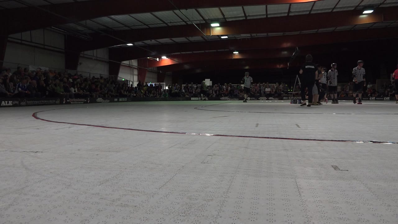 Low rink-level view with referees, skaters, and spectators indoors.