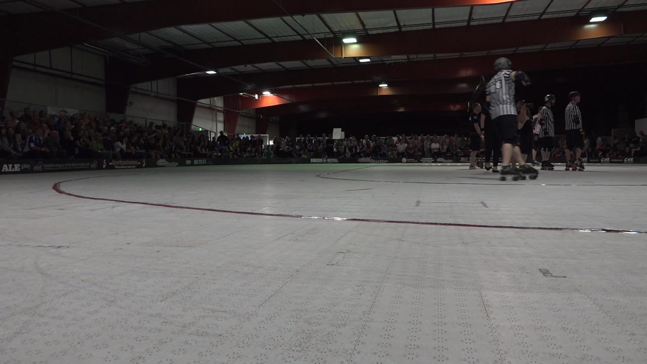 Low floor-level view across a white roller derby rink in Denver, Colorado, with a red curve, striped referees near the right side, skaters, and seated spectators behind them.