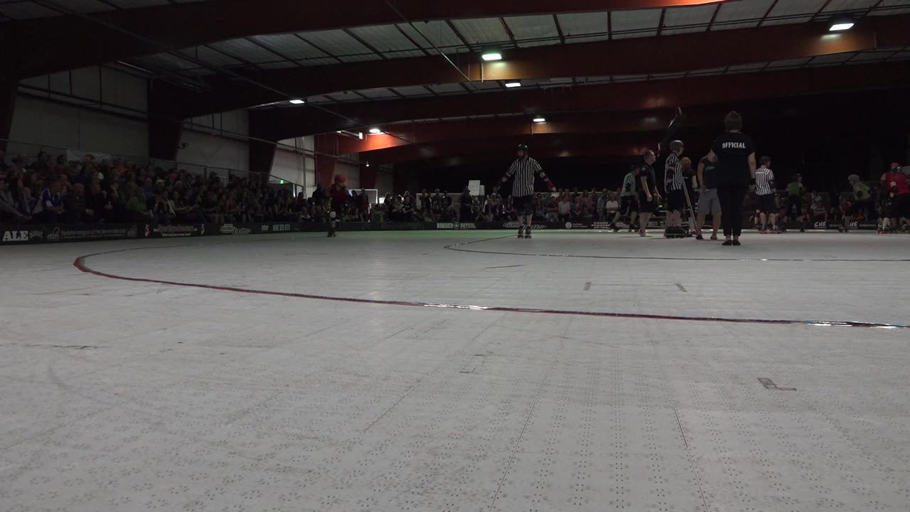 Low ground-level view of a Denver indoor roller rink in 2015, with skaters and officials gathered near the far side and a seated crowd in the background.