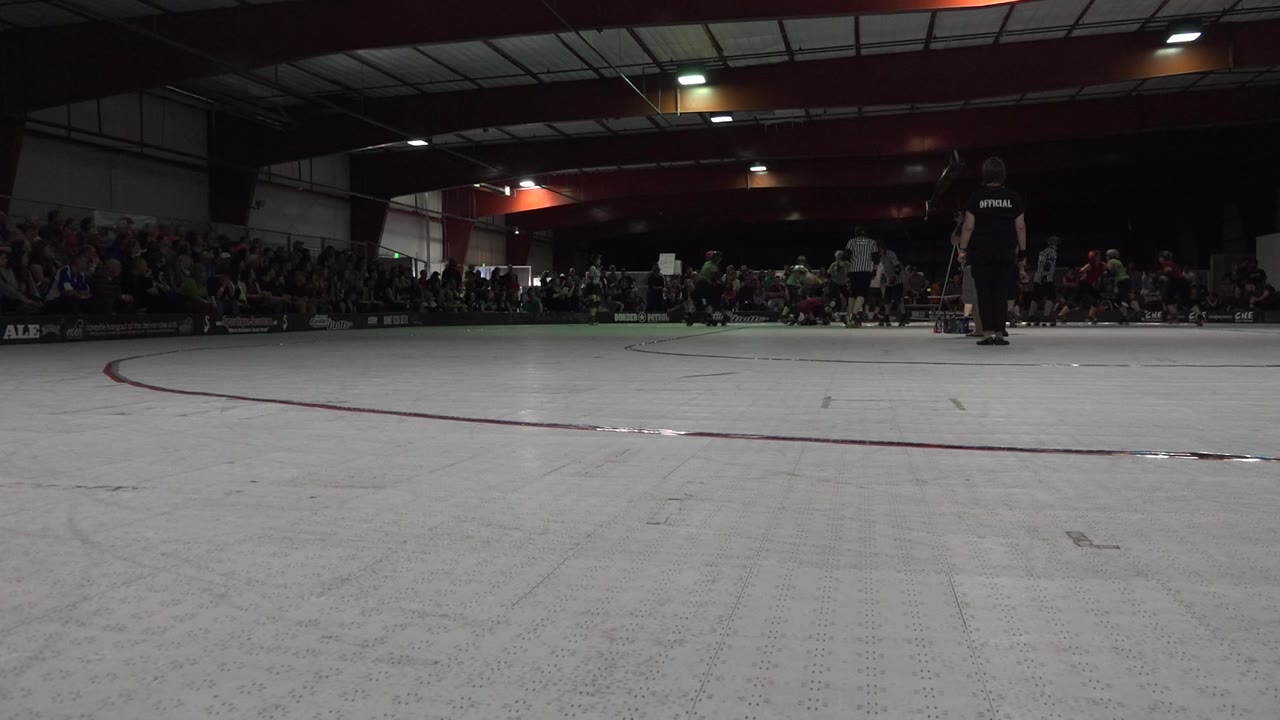 At 0:00, a low Denver roller rink view shows a pale floor in the foreground, skaters near the center, officials to the right, and a seated crowd along the back wall.