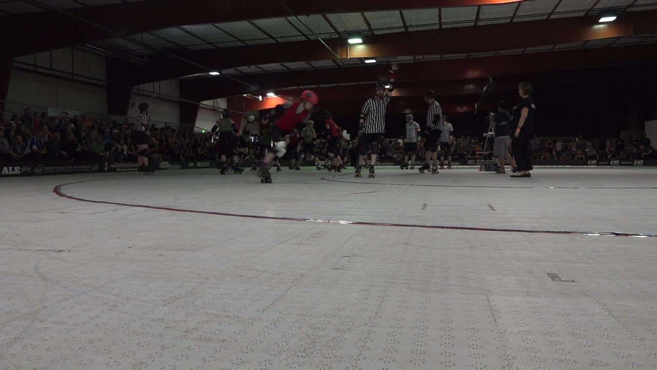Skaters in helmets move across the Denver rink as striped referees stand near the middle of the frame, with seated spectators along the back wall and overhead lights reflecting on the floor.