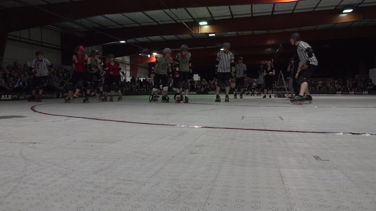 Low rink-level view in Denver, Colorado, showing roller derby skaters in helmets and referees in striped shirts gathered near the far side with spectators in the background.