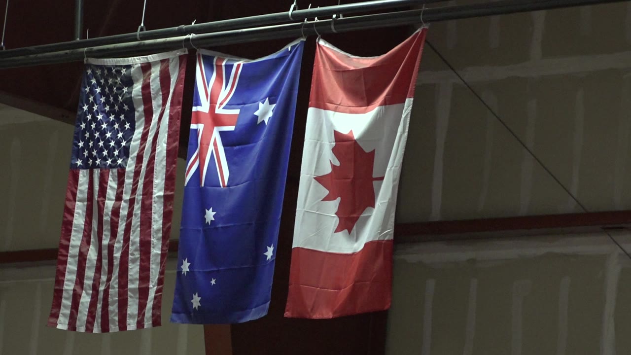 USA, Australia, and Canada flags remain centered under the ceiling rail in the Denver venue, with the wall panels and beam visible behind them and no visible people.