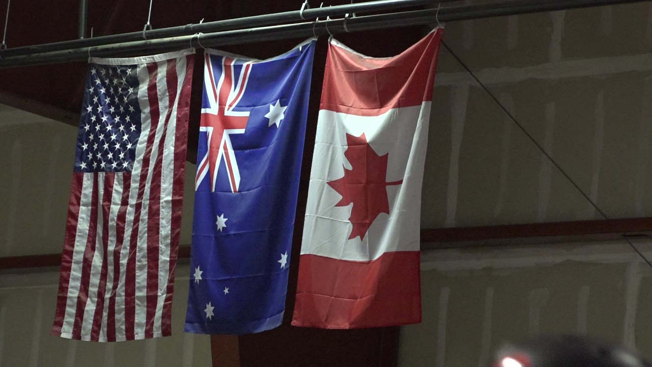 Three flags, United States, Australia, and Canada, hang from a pipe above a school-stage wall in Denver, with no visible people in this opening frame.