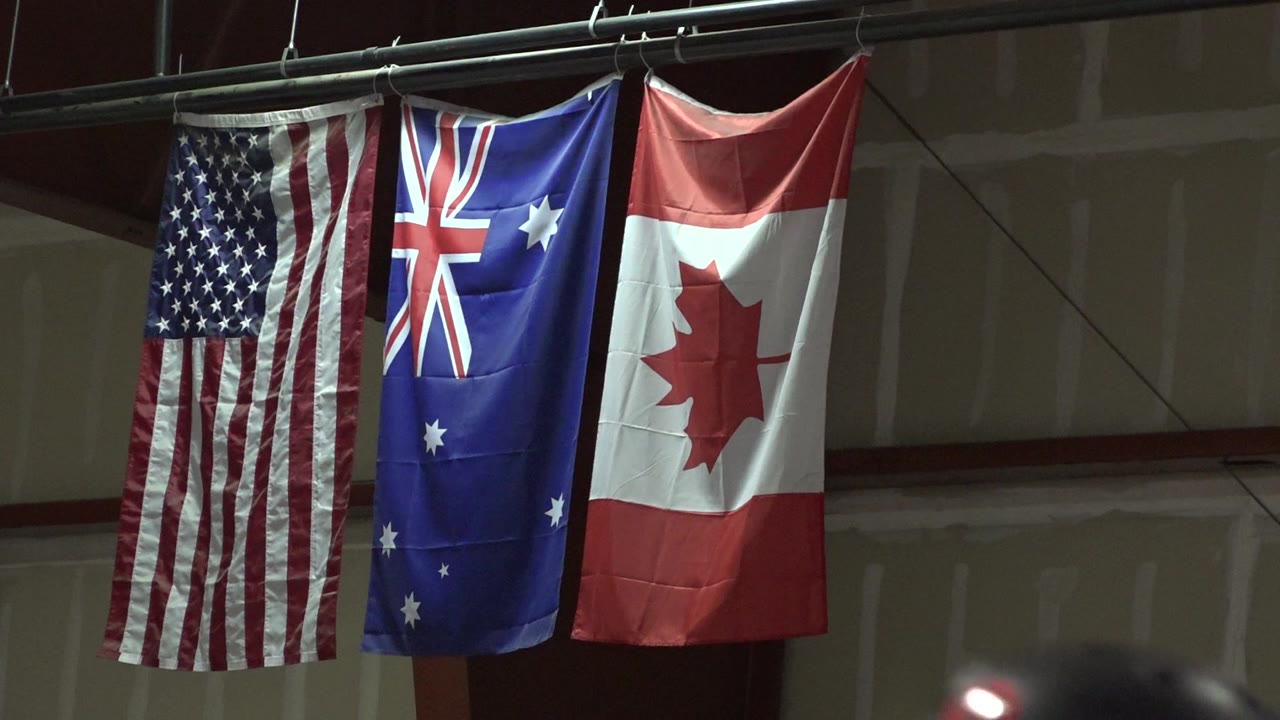 The Denver school-stage view holds on the three hanging flags, with the United States flag at left, Australian flag in the middle, and Canadian flag at right.