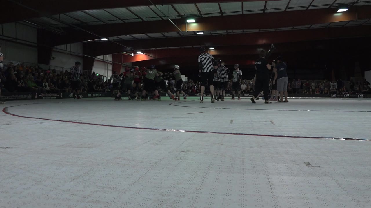 At 0:01, the low camera frames a cluster of derby skaters and referees across the rink, with the crowd still seated along the back wall of the Denver venue.