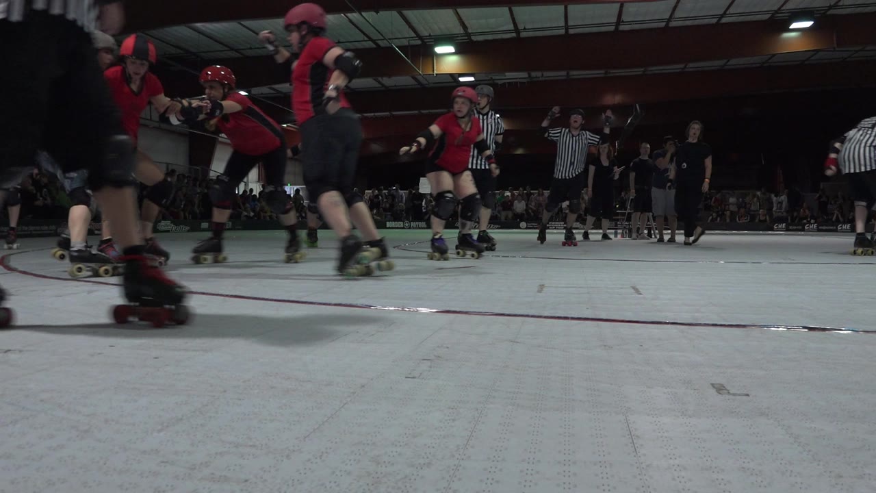 Denver roller derby skaters in red and dark uniforms pass close to a low camera, with referees and spectators visible in the background.