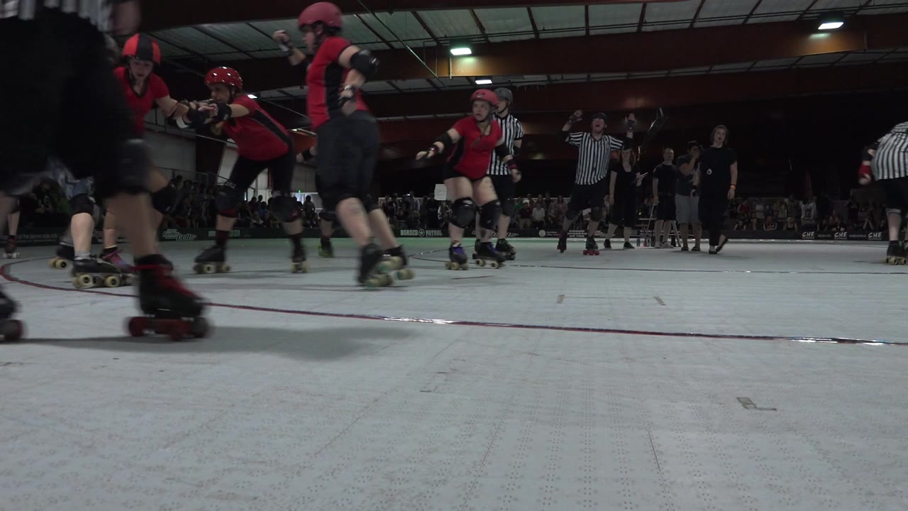 A green-uniformed skater faces the pack as red-uniformed skaters roll across the Denver indoor derby track, with the crowd behind them.