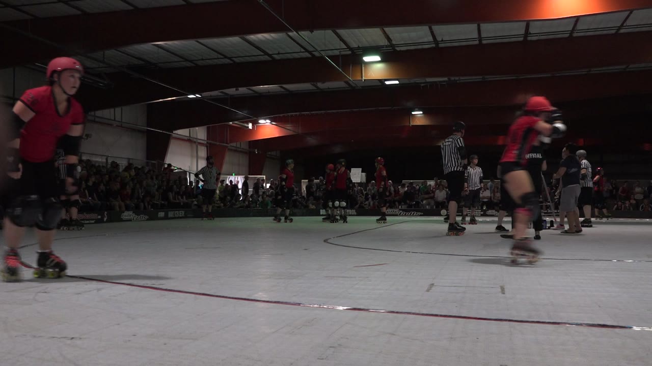 Skaters in protective gear pass across an indoor rink.