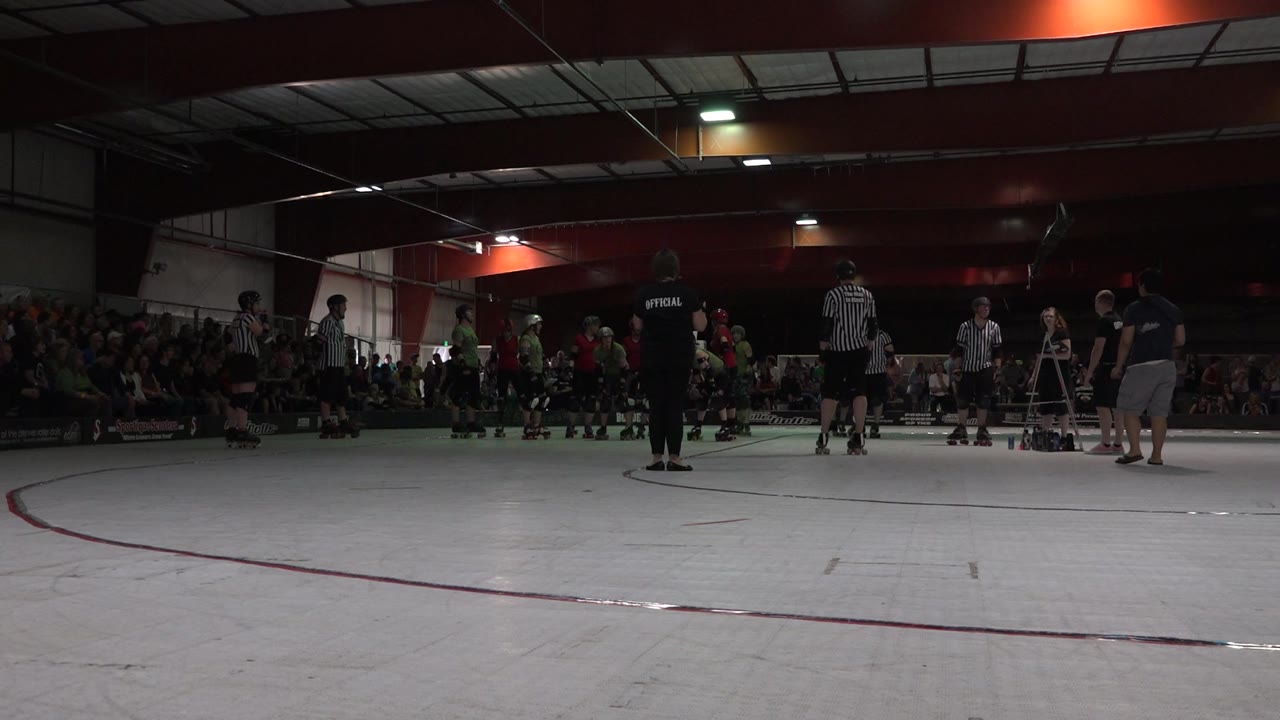 Denver indoor rink frame with a black-shirted official centered, striped referees nearby, players in green to the left, and a seated crowd behind them.