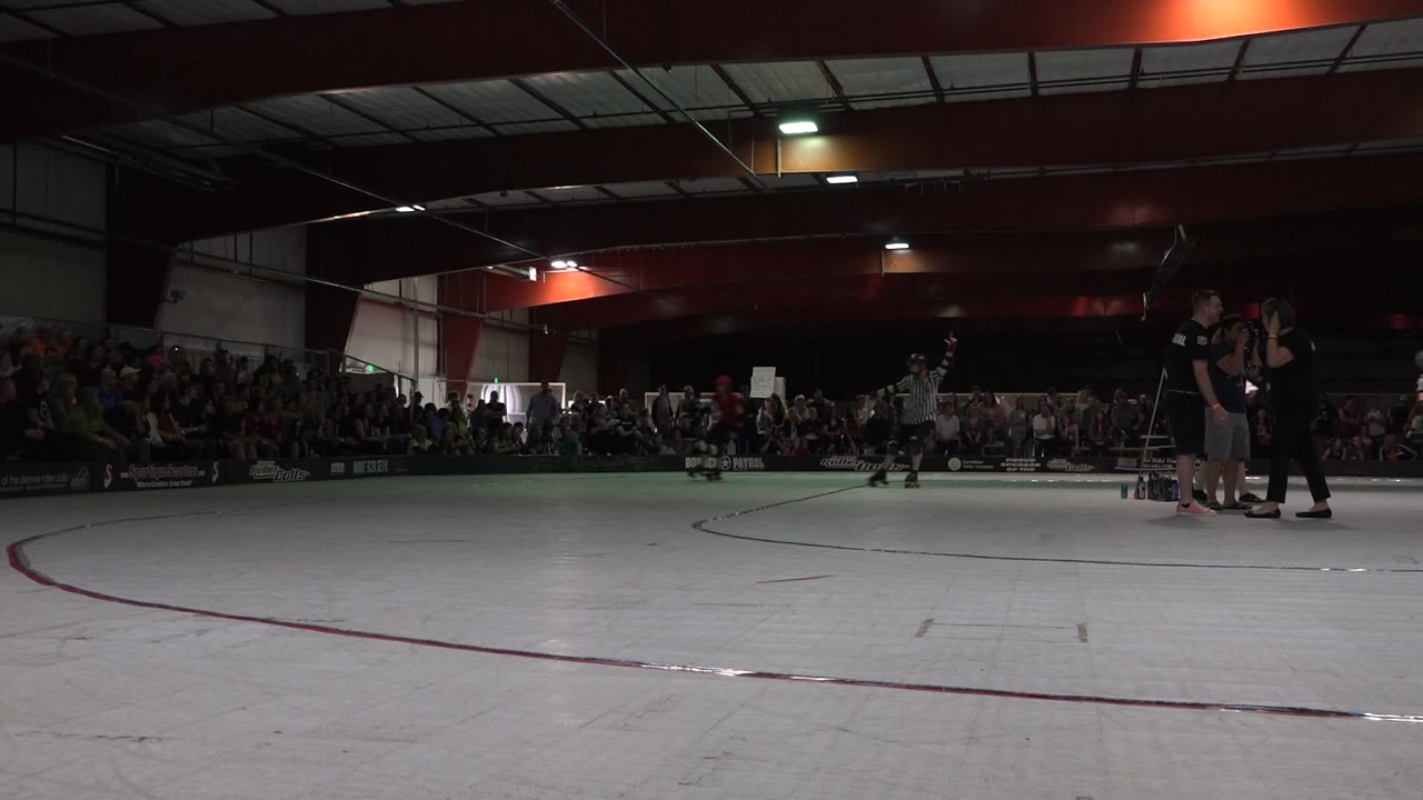 Wide Denver indoor roller rink view with a seated crowd in the background, officials and staff near the far side, and open rink floor in front.