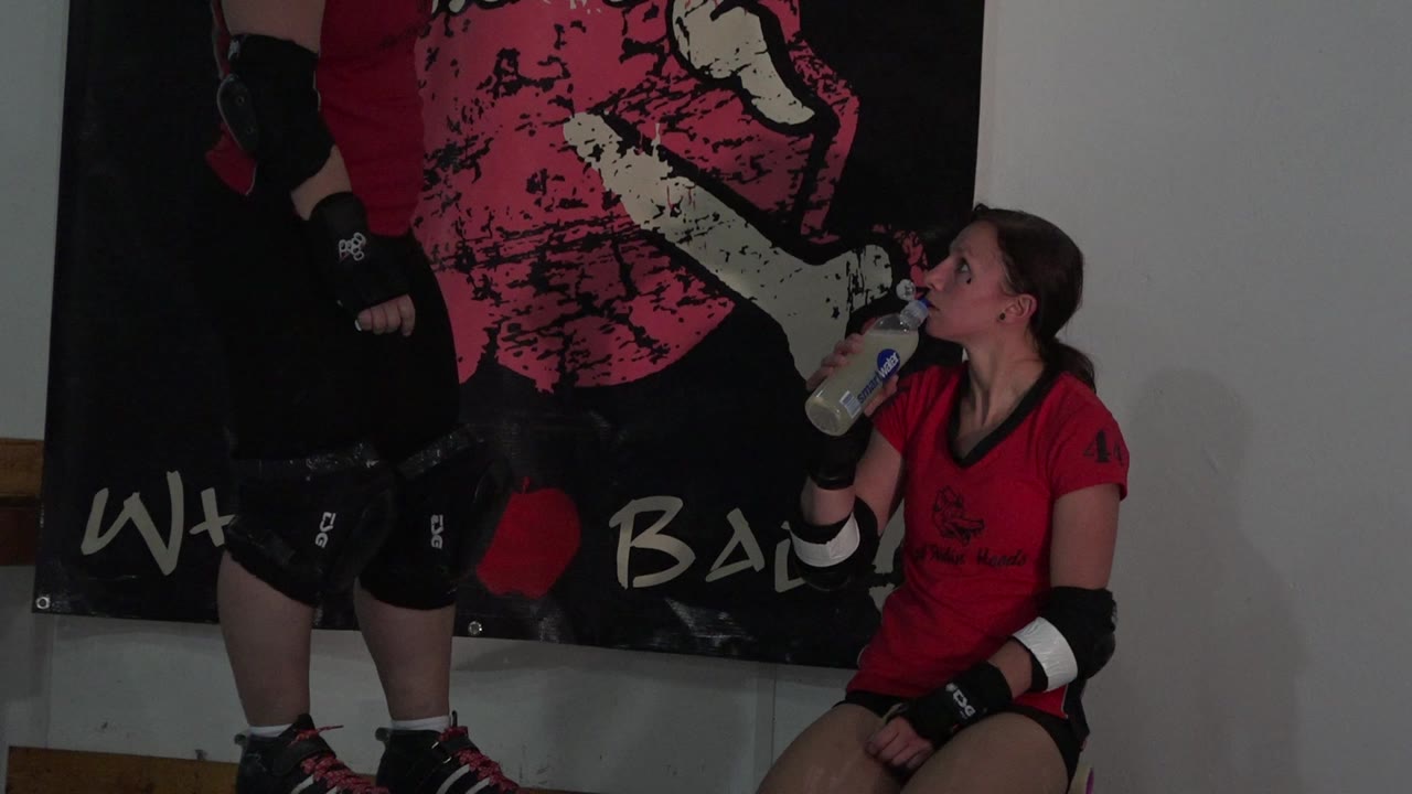 Two red-uniformed skaters beside a wall banner, one seated drinking water.