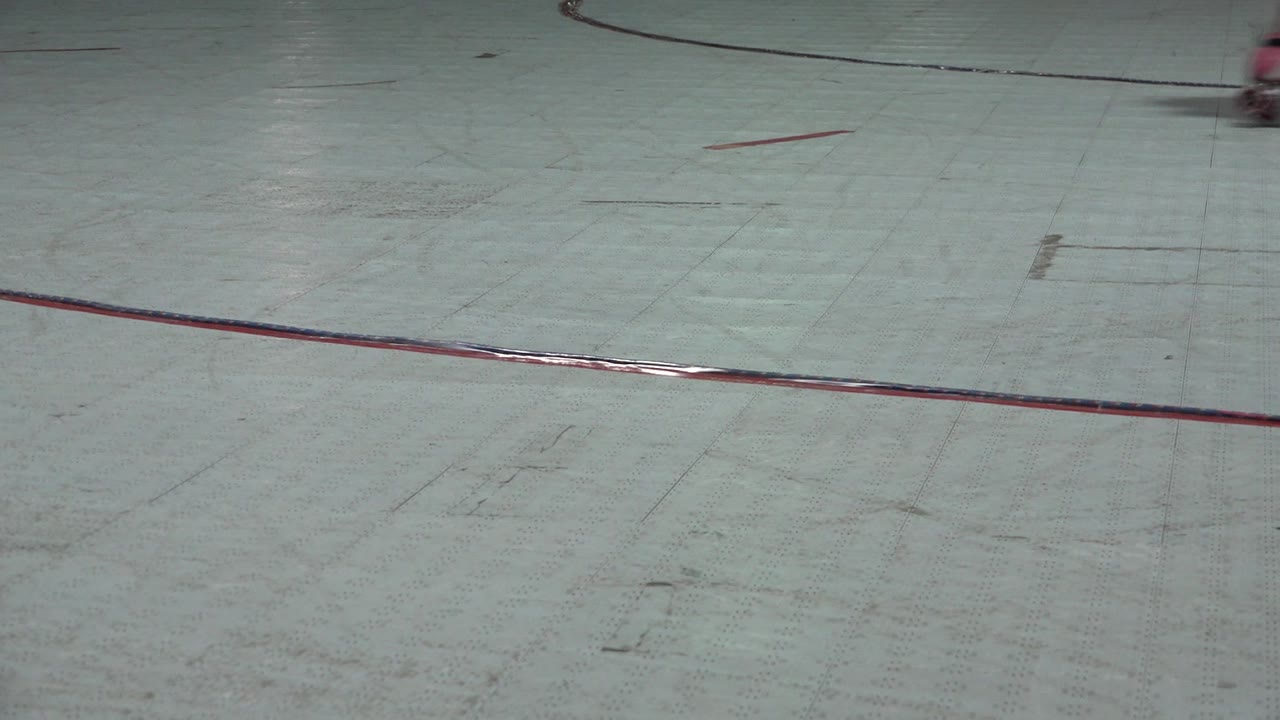 Empty pale skating floor with red rink lines and distant cropped skaters.
