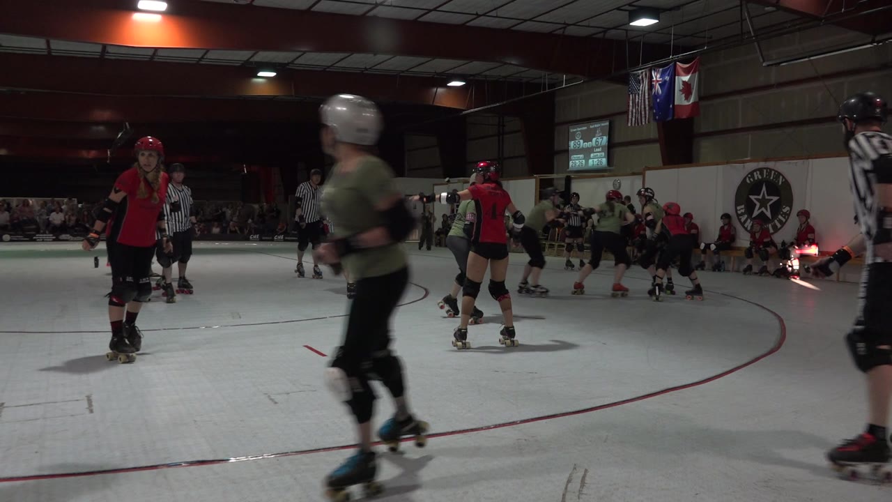 A skater blurs through the foreground while a red-shirted derby player and referees stand on the track in the Denver indoor arena.