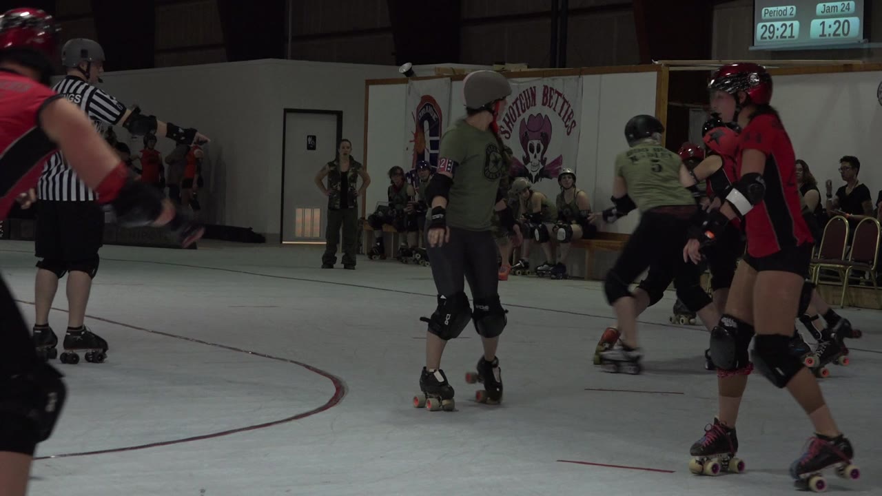 Skaters in helmets and pads cluster near the inside line of the Denver indoor rink while a referee stands in the foreground and spectators watch from the back wall.