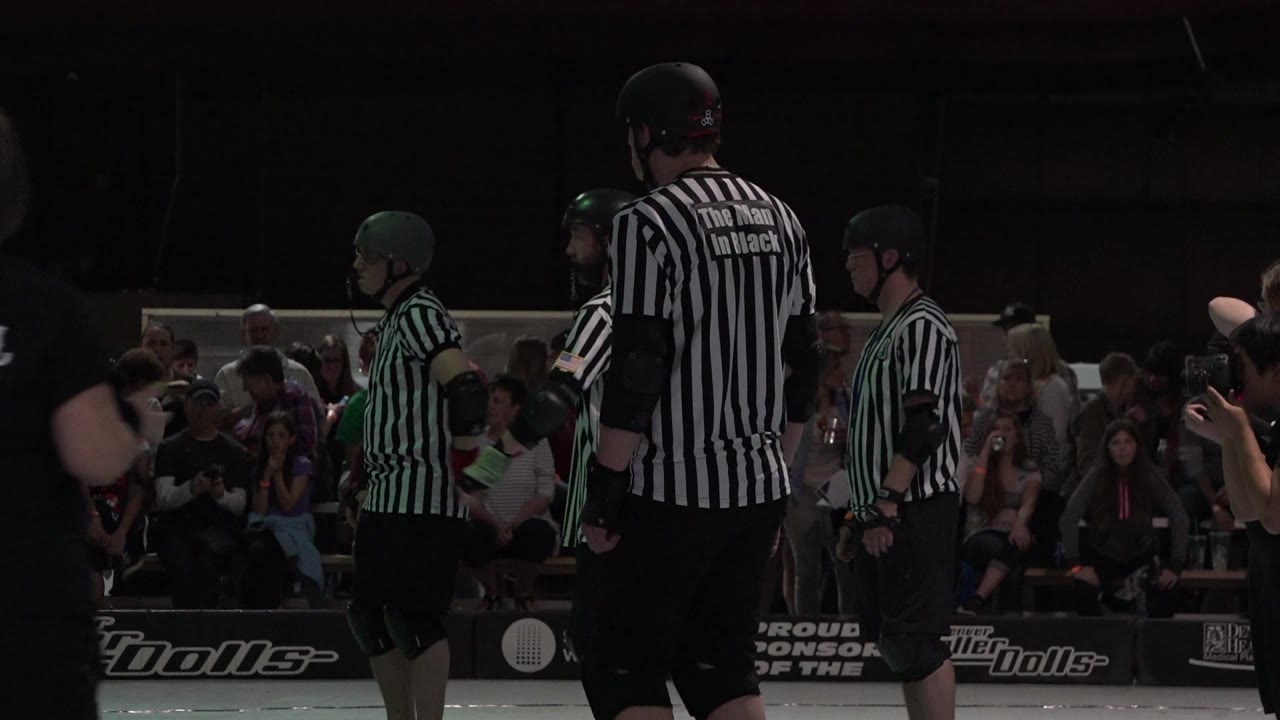 Striped roller officials and helmeted participants stand in front of spectators.