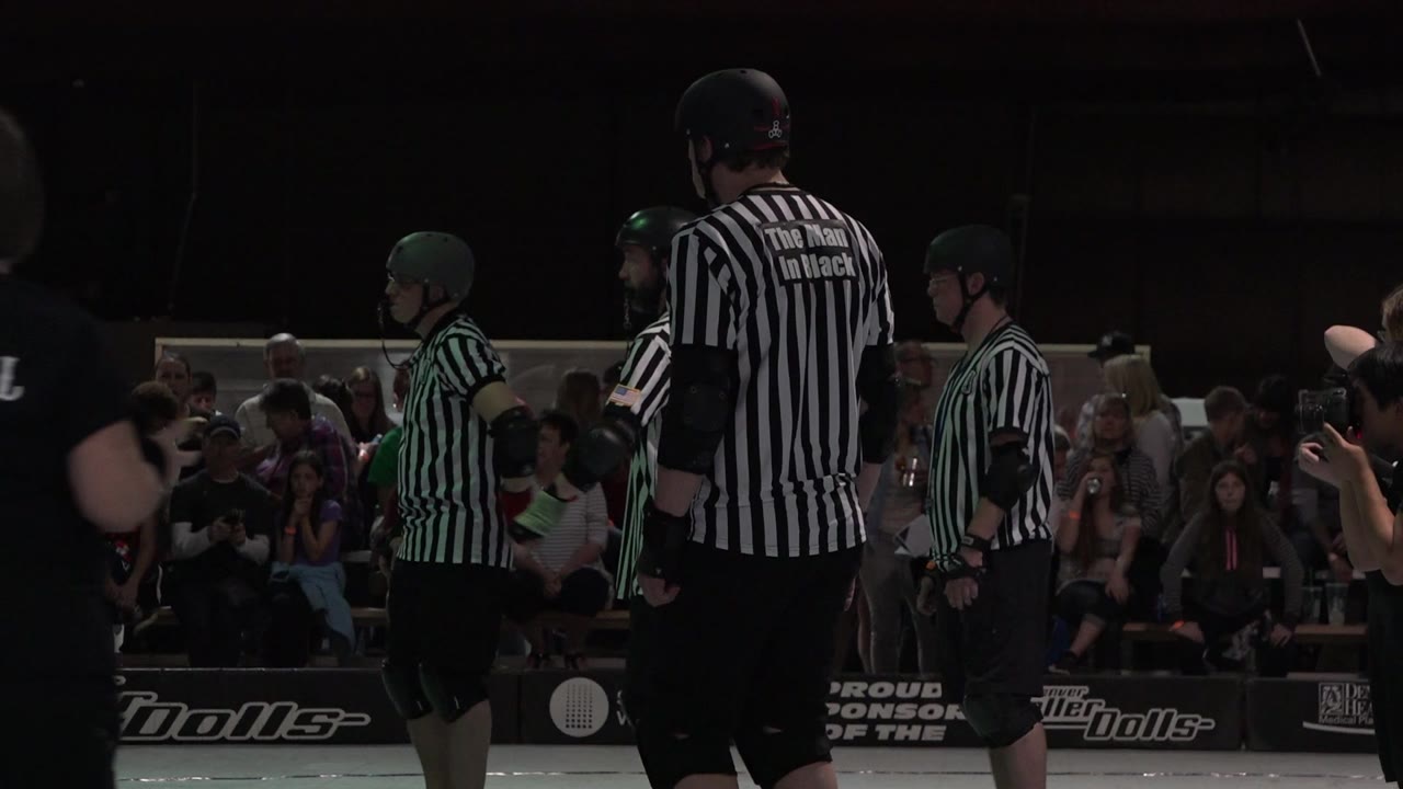 Denver roller event frame with a tall striped official in the foreground, helmeted skaters beside him, and spectators watching from behind the track barrier.