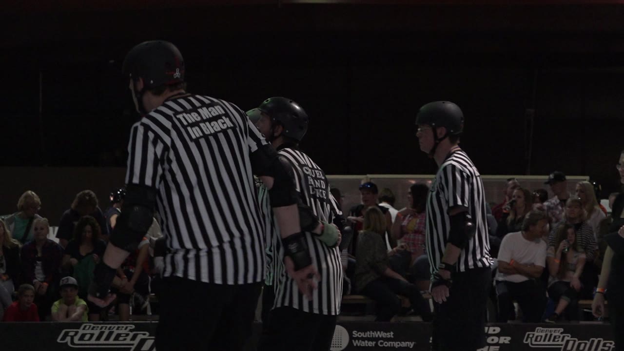 Roller derby referees in striped shirts standing by an indoor rink.