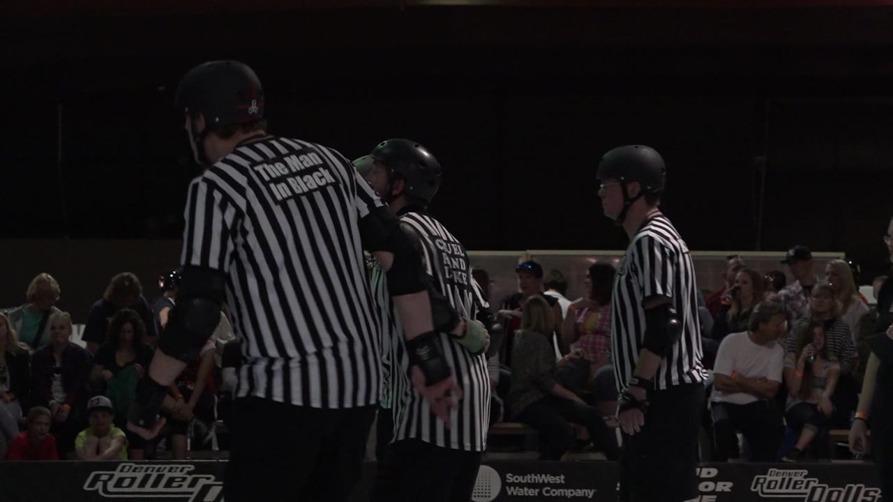 A tall referee with a black helmet stands in the foreground at a Denver roller derby rink, with other officials and seated spectators behind the barrier.
